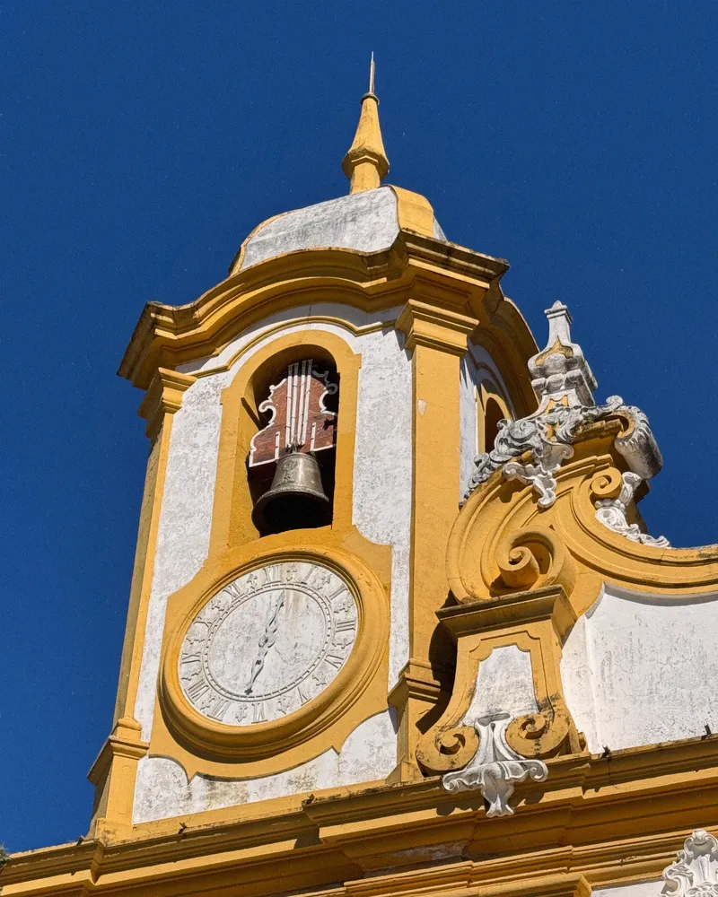 Torre de igreja barroca amarela e branca, com sino e relógio, contra um céu azul intenso.
