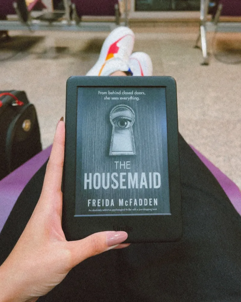 Kindle com o livro The Housemaid, de Freida McFadden, na sala de embarque de um aeroporto.