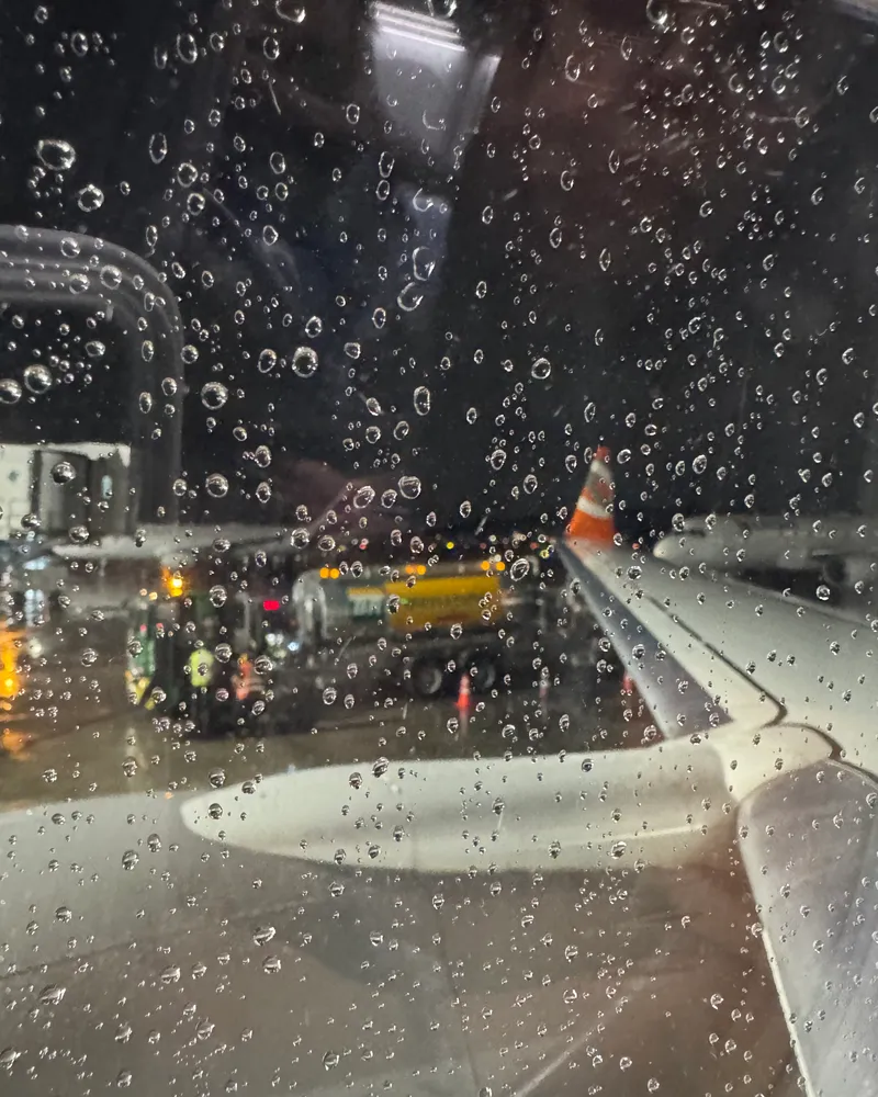Vista da janela do avião com gotas de chuva, a asa e a pista do aeroporto à noite.
