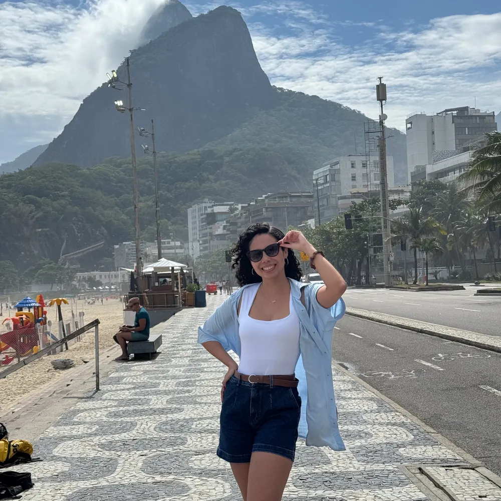 Sandrele sorrindo de óculos escuros no calçadão da orla do Rio de Janeiro, com o Morro Dois Irmãos ao fundo.